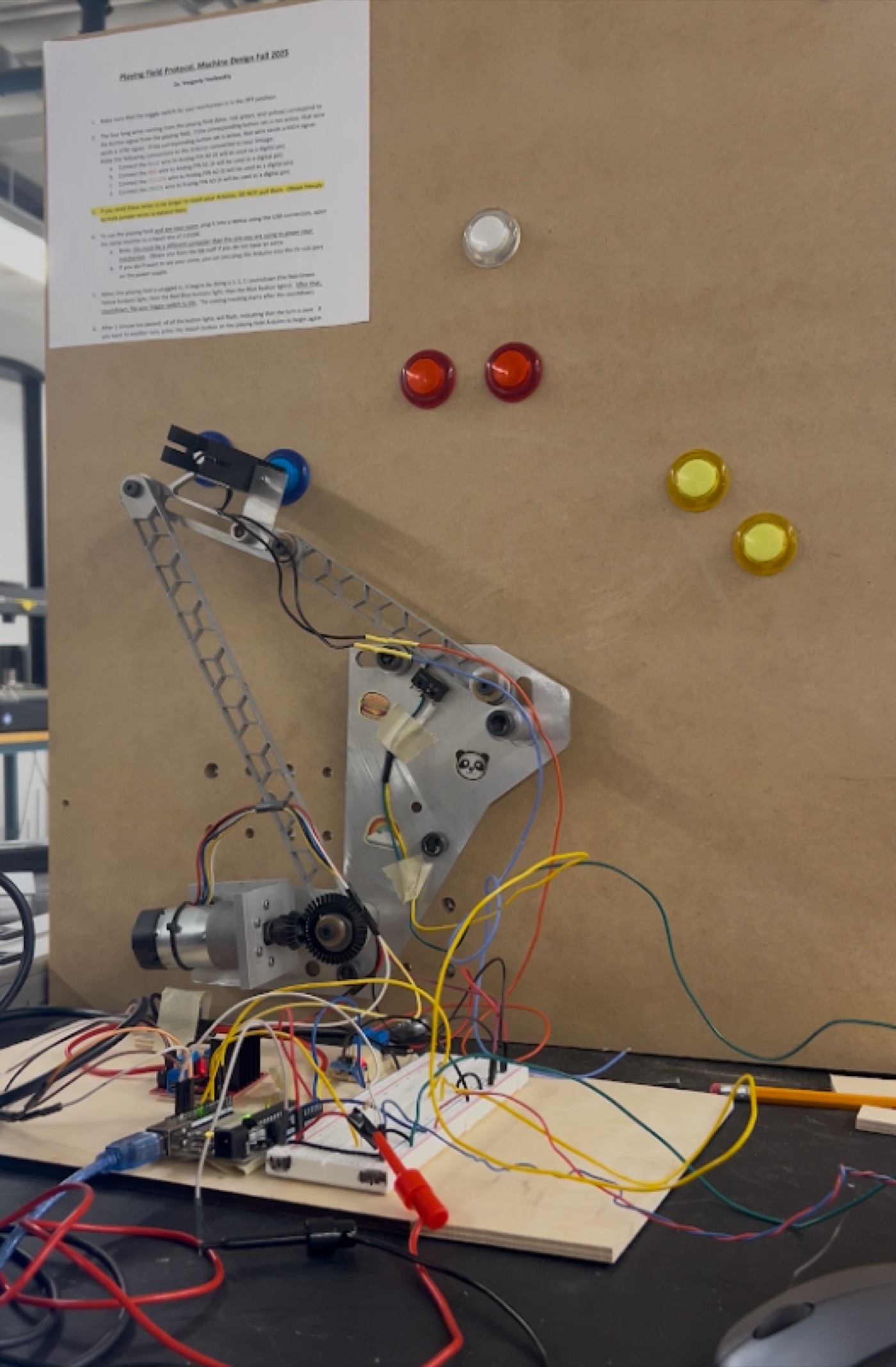 The four-bar linkage mechanism mounted on a wall board next to the game board's colored buttons, wired to an Arduino and breadboard.